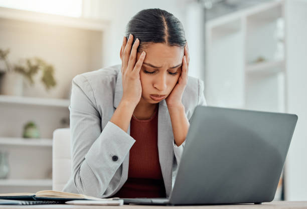 A young Hispanic woman is sitting at her desk in her office. She has her laptop open in front of her and she is holding her head with her hands.