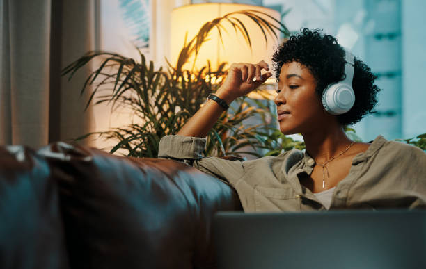 A young African American woman is sitting on the couch of her living room. She has her laptop open in front of her and is wearing headphones.