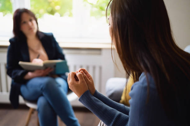 An Asian American woman is sitting in a therapy office across from her therapist. The therapist is holding a booklet in her hands as she writes.