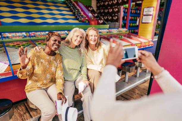 Three senior citizen women are sitting beside each other getting their picture taken. Behind them is a carnival game. They are all smiling with their arms around each other.
