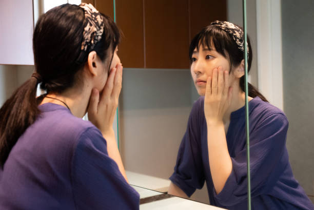 A young Asian American woman is standing in her bathroom, looking at herself in the mirror. She is holding her hand up to her cheek with a blank stare.