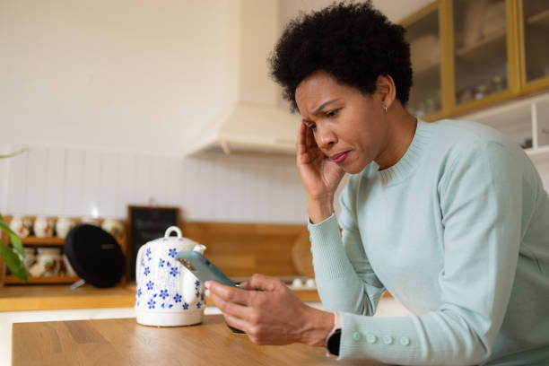 An African American woman is standing in the kitchen of her home. She is leaning on the counter with her hand on her head, as she looks at something on her phone.