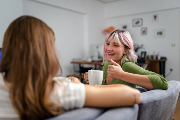 Two young women are sitting on the couch in a living room. They are both talking and smiling together. One of the women is holding a coffee mug in her hand.