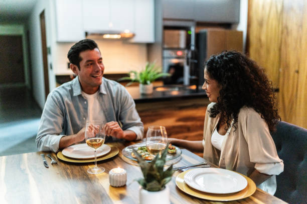 An interracial couple is sitting at their dining table having dinner. They have glasses of wine in front of them. They are both smiling as they talk.