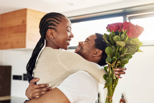 A young African American couple is together in their kitchen. The man is carrying the woman as she holds red roses in her hand.