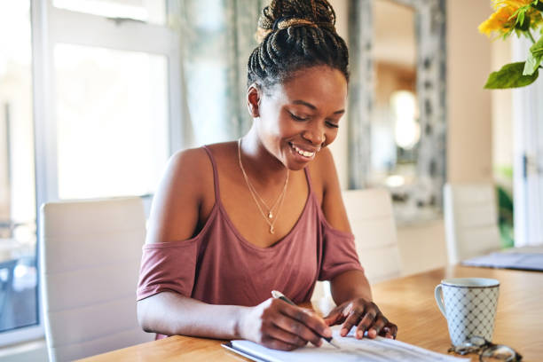A young African American woman is sitting at her dining table working. She is holding a pen in her hand as she writes on a piece of paper. There is a coffee cup placed next to her on the table.