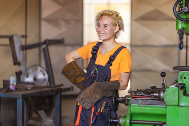 A young blonde woman is standing in her work place at a mechanic shop. She is wearing work gear and gloves. This woman is actively at work.