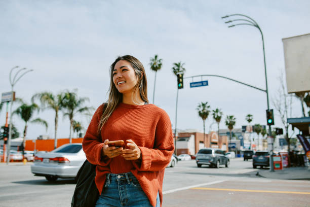 A young Asian American woman is crossing the street in Los Angeles. She is holding her phone in her hand and is smiling.