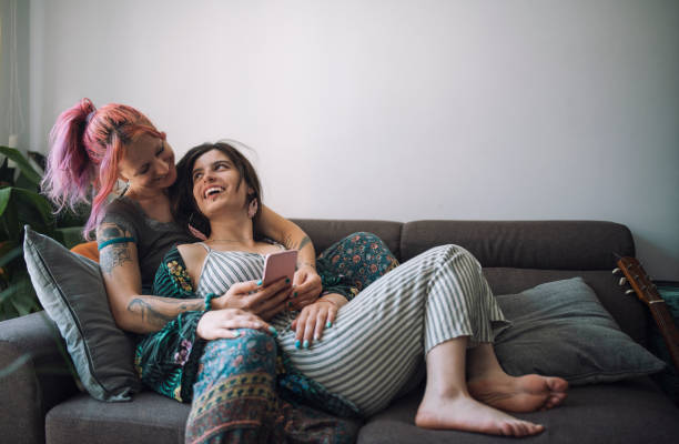 A young lesbian couple is laying together on the couch of their living room. They are looking at each other and smiling as one of them holds a phone in their hand.
