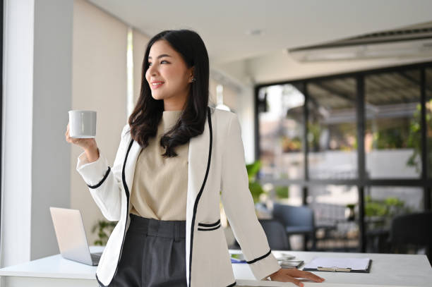 A young Asian American woman is standing in her office. She is holding a coffee cup and smiling. Woman is at work.