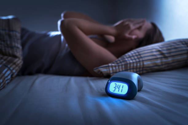 A young woman is laying in her bed in the dark. She has her hands covering her face, as her alarm clock displays 3:41 am.