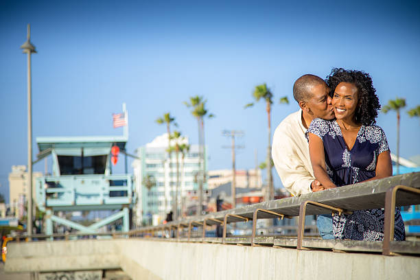 An African American couple is standing on the pier at the beach.The man is kissing the woman on the cheek, as he hugs her from the back.