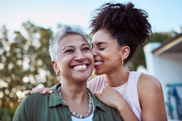A biracial mother and daughter are standing outside together. The daughter is hugging her mother from the back as she nuzzles her face onto her mothers. They are both smiling.