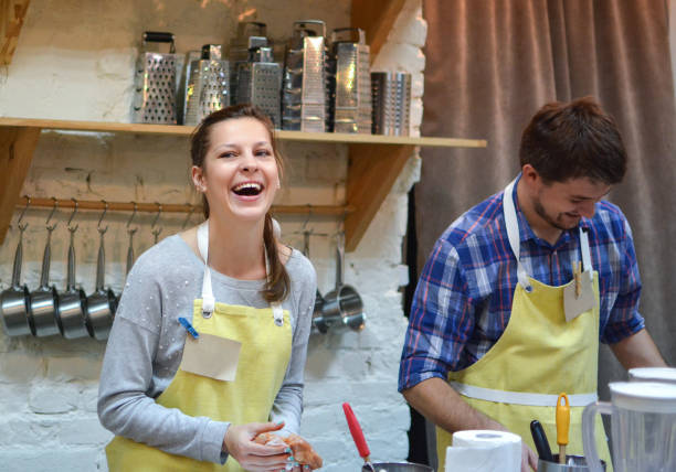 A young couple is taking a cooking class together. They are standing beside each other with yellow aprons on as they laugh together.