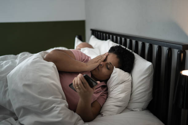 An African American woman is laying in bed beside her husband. She has her eyes closed as she puts her hand on her face and is holding her phone in her hand.
