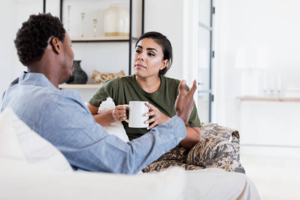 A young biracial couple is sitting together on their couch. They are facing each other as they talk. The young Hispanic woman is holding a cup of coffee in her hand.