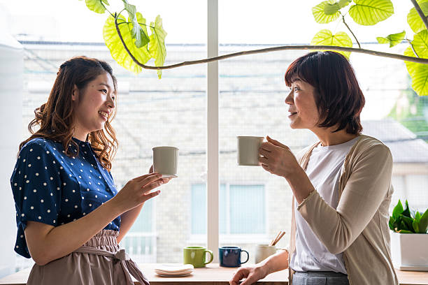 An Asian mother and daughter are standing together in their kitchen in front of each other. They are both holding coffee mugs in their hands. Set Boundaries with Your Mother