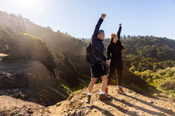 A young couple is on a hike together. They are both raising their hands as they yell and smile together. The couple is holding hands as well.