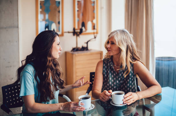 A mother and daughter are sitting together at their dining table. They are both talking to each other as they drink coffee. Set Boundaries with Your Mother