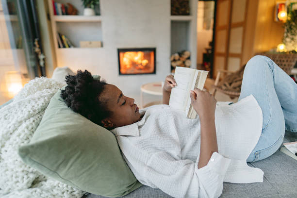 A young African American woman is laying on her couch with her legs up. She is reading a book that is resting on her chest.