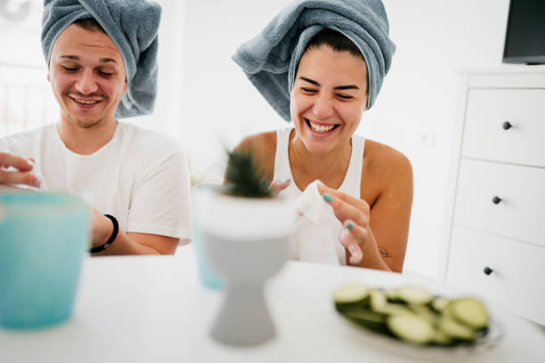 A young couple is doing a spa day together. They both have towels wrapped around their heads as they put cucumbers on their eyes.