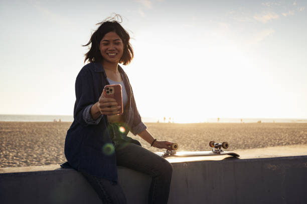 An Asian American woman is at the beach sitting down next to her skateboard. She is smiling as she looks at her phone in her hand.