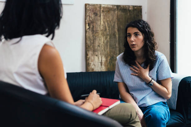 A young woman is sitting across from her therapist in her office. She is holding her hand on her chest. The therapist has a notebook on her lap.