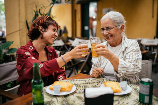 A mother and daughter are sitting together at a restaurant. They are both eating food and doing a cheers with their glasses of beer. 
