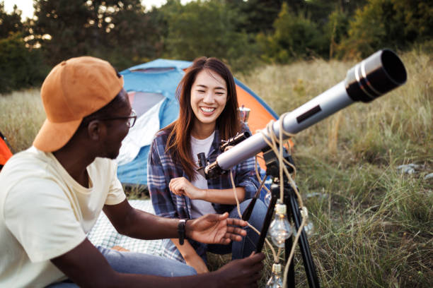 A biracial couple is camping together in the outdoors. They are stargazing with a telescope. Their tent is behind them.