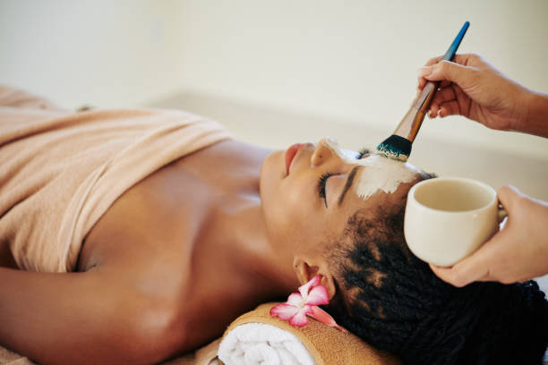 A young African American woman is at a spa laying down. She is wrapped in a blanket as she has a face mask being put on her face.