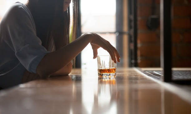A woman is sitting at a bar with a glass of whiskey in front of her. She is holding the glass by the rim.
