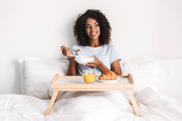 A young African American woman is laying in her bed with a table in front of her. She is eating oatmeal as she smiles. On the table there is a croissant and a glass of orange juice.