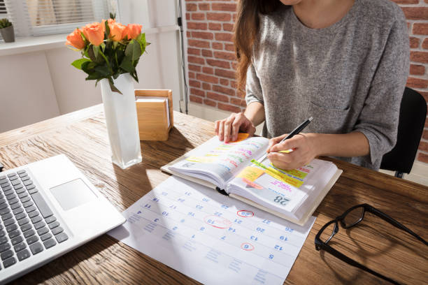 In this image there is a a planner and calendar on a desk. A woman is writing in her planner. Her laptop is open in front of her.