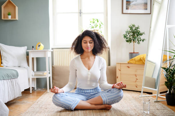 An African American young woman is meditating on the floor of her bedroom. She has her legs crossed with her eyes closed.