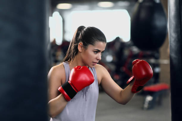A young woman is at the gym wearing red boxing gloves as she punches a punching bag.