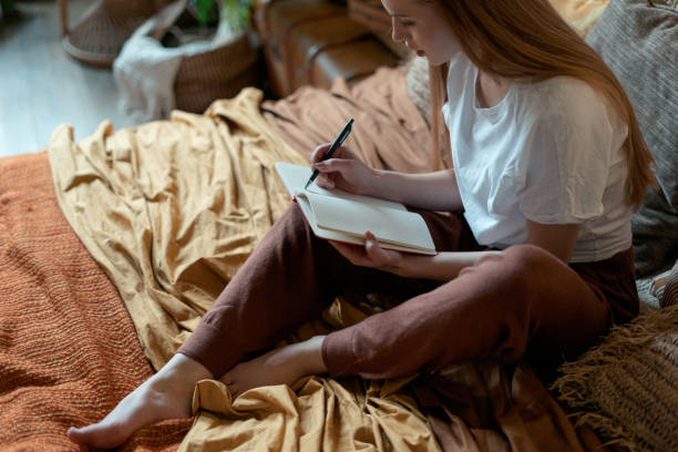 A young American woman is laying on top of her bed. She has her notebook open as she writes in it.