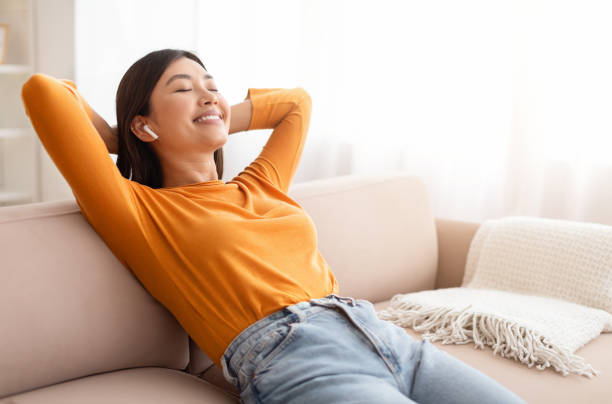 A young Asian American woman is relaxing on her couch wearing an orange long sleeve shirt. She has her hands behind her head as she listens to music on her earphones.
