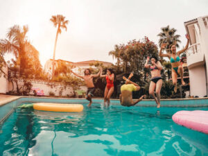 A group of four young friends are having a pool party. They are all jumping in the pool together at the same time. There are two floaties in the pool.