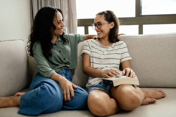 A mother and daughter are sitting beside each other on the couch with their legs up. The daughter has a book on her lap as they both smile at each other.