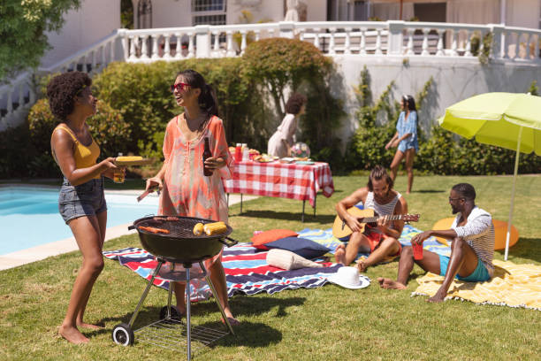 A group of friends is having a pool party in their backyard. Two of the women are standing around the grill as they talk together. Two men are sitting on the floor on blankets as one of them plays the guitar.