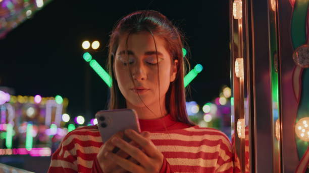 A young teenage girl is at a carnival looking at her phone. She has a somber facial expression.