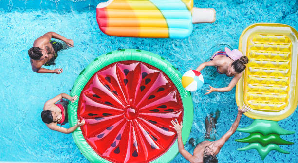 In this image there are four friends in a pool throwing a beach ball to each other. Surrounding them in the pool are three floaties, one pineapple, one watermelon, and one popsicle.