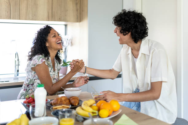 A Hispanic mother and son are sitting together in the kitchen. There is breakfast food on the table as they hold each others hands and laugh together.