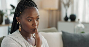 A young African American woman is sitting in her living room. She has a blank stare as she bites her finger nail.