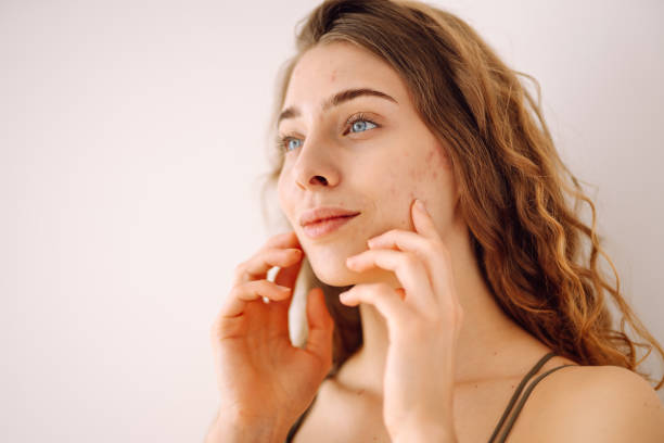 A young woman is smiling as she looks in the distance. Behind her is a white background. She has her hands on her face.