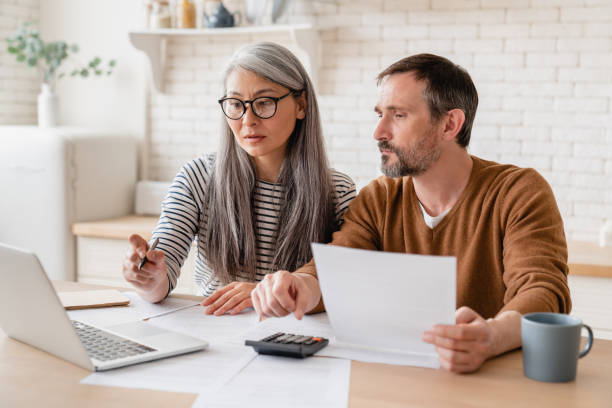 Mature middle-aged couple family wife and husband counting funds, savings declarations, investments, paperwork, financial documents, bankruptcy, court case, bills, pension with laptop.