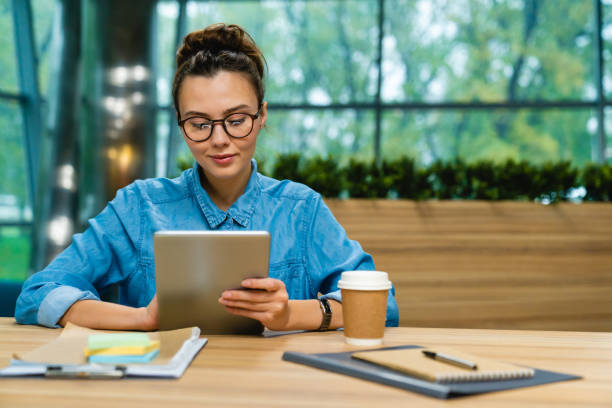 A woman is sitting at a cafe doing work. In front of her on the table are notebooks and pens, and a coffee cup. She is doing work on her tablet.