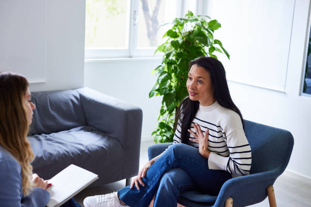 A young woman is sitting in her therapists office. She has her legs crossed and her hand on her chest as she speaks. The therapist is sitting in front of her with a notepad.