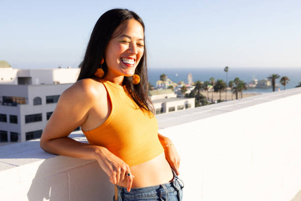 A young Asian woman is standing against the ledge of a rooftop. In the distance there is the beach. She is smiling as the sun shines on her face.