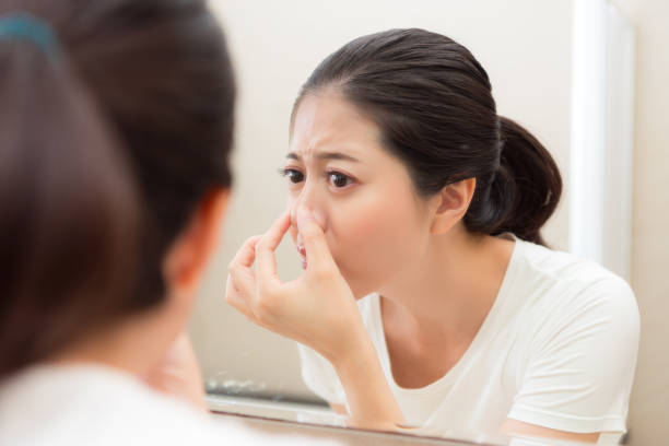 An Asian American woman is looking at herself closely in the mirror. She is pinching her nose as she leans into the mirror.